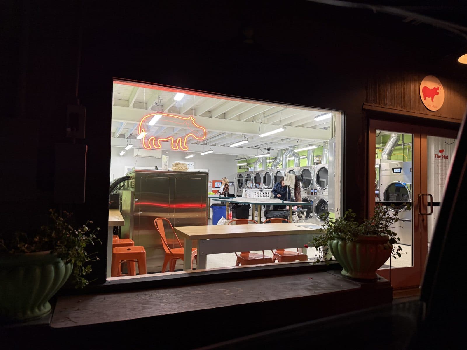 The Mat at night, seen from outside, with the lit interior and neon hippo sign visible through the window