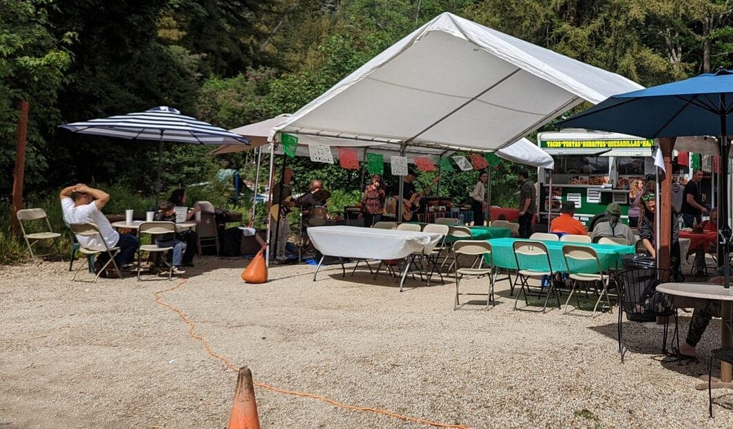 The El Rey Leon taco truck parked under the trees in the lot at The Mat, with tables and umbrellas set up beside it